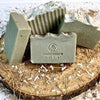 Four handmade greenish-gray soap bars with textured tops and natural speckles, one stamped with "HANDMADE Natural," arranged on a wood slice with dried herb shavings, featured in Health & Wellness, Soaps, and Best Sellers collections.