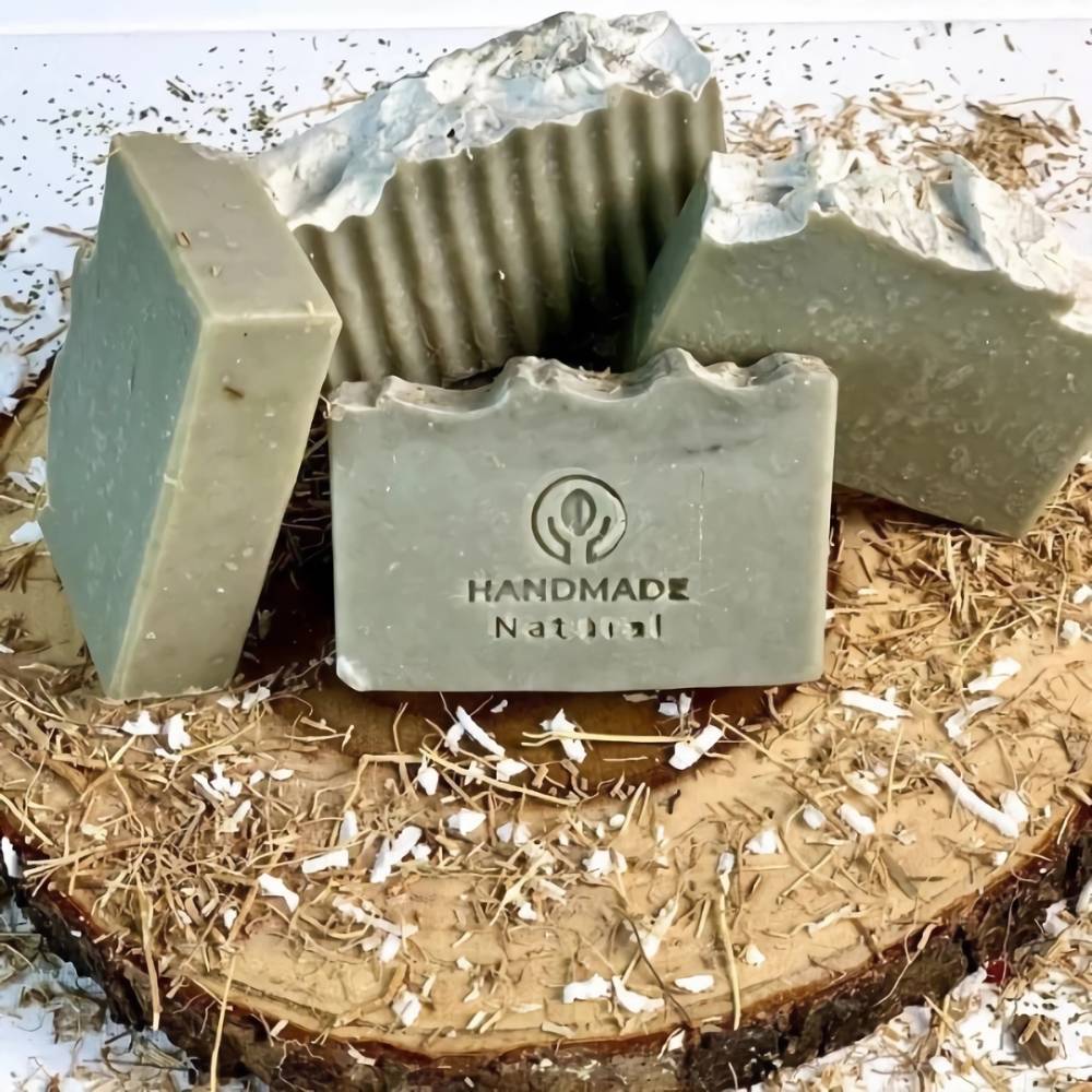 Four handmade greenish-gray soap bars with textured tops and natural speckles, one stamped with "HANDMADE Natural," arranged on a wood slice with dried herb shavings, featured in Health & Wellness, Soaps, and Best Sellers collections.