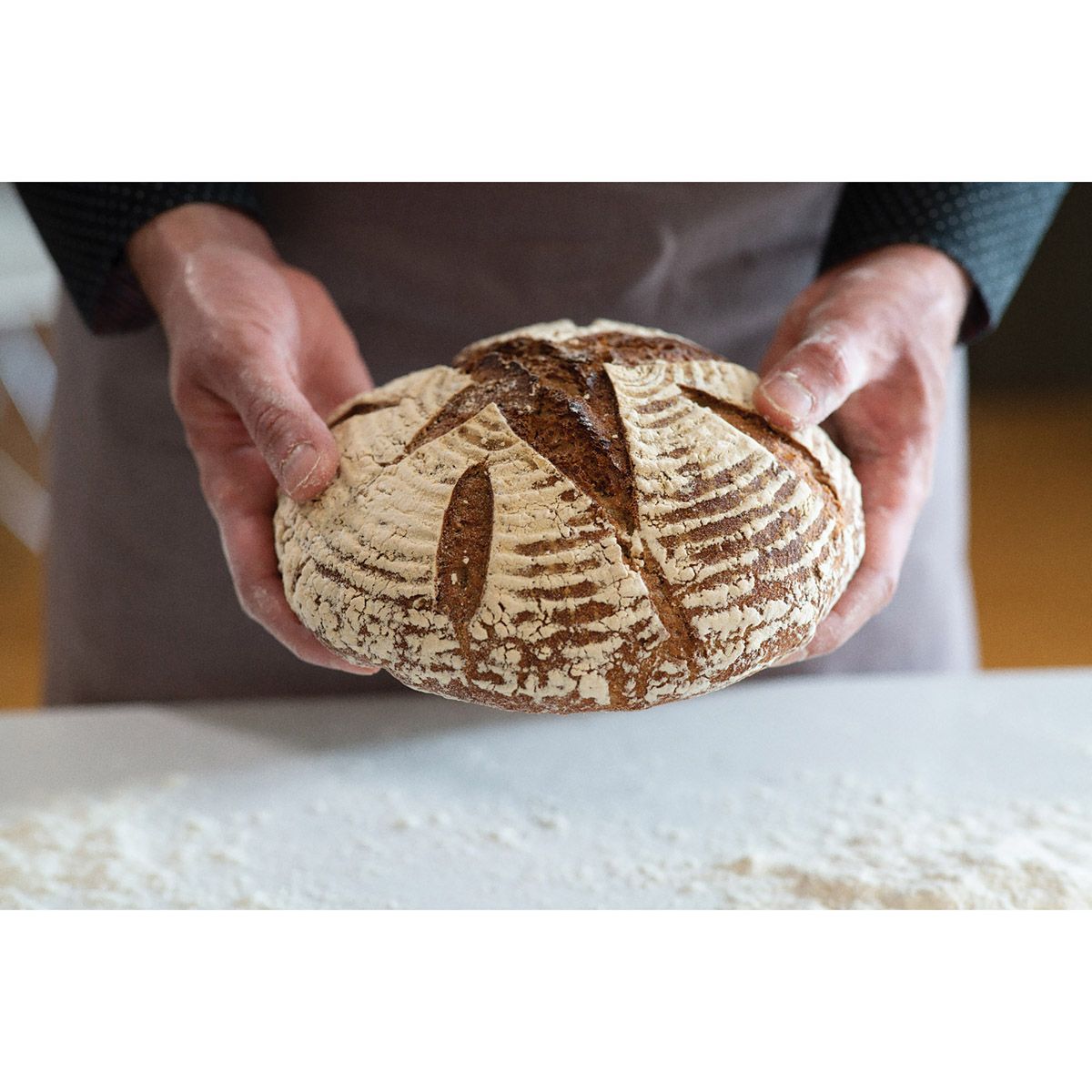 Hands gently holding a rustic artisan sourdough loaf with a patterned crust, set against a floured surface, highlighting homemade bread baking in Kitchen & Food and Baking Supplies.