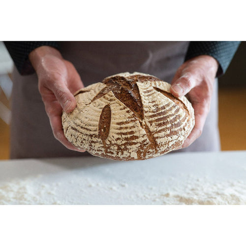 Hands gently holding a rustic artisan sourdough loaf with a patterned crust, set against a floured surface, highlighting homemade bread baking in Kitchen & Food and Baking Supplies.