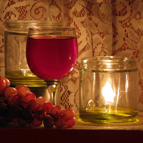 Handcrafted oil lamp burning with olive oil in a clear glass jar, accompanied by a wine glass filled with red liquid and a cluster of purple grapes, featured in Lamps & Lighting, USA Made, Oil Lamps, Olive Oil Lamps, and Best Sellers collections.