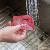Hand holding a red polycarbonate pan scraper under running water in a kitchen sink, ideal for Kitchen & Food, Cookware, Cast Iron, Best Sellers, and Promo Eligible collections.