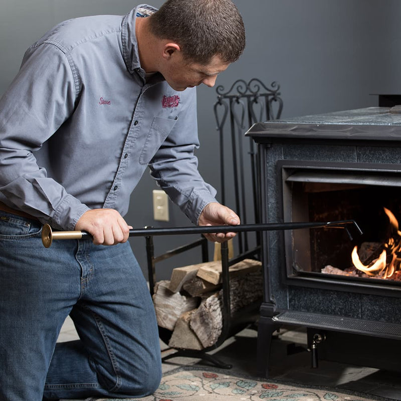 Man kneeling while using a handcrafted powder-coated steel fire tool with brass mouthpiece to tend logs inside a cast iron stove, ideal for Hearth Accessories and Stoves collections
