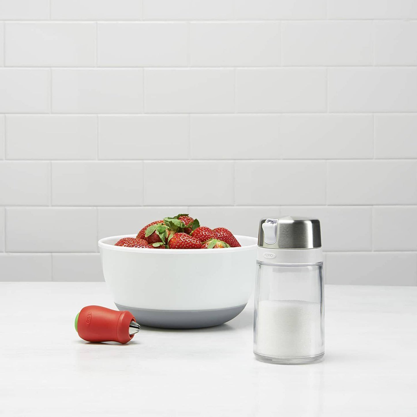 Clear glass sugar dispenser with stainless steel lid and side spout for controlled pouring, designed to prevent clumping, shown next to a white bowl of fresh strawberries, ideal for Kitchen & Food, Kitchen Gadgets, Kitchen Accessories, Storage and Serving, Best Sellers, and Promo Eligible collections.
