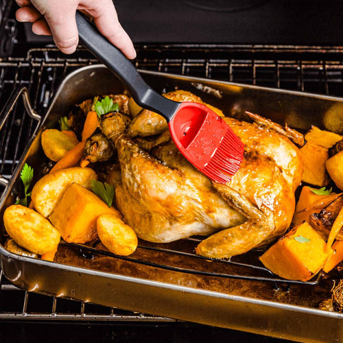 Red silicone basting brush with ergonomic black handle applying glaze to roasted chicken and vegetables in oven-safe pan, featured in Kitchen & Food, Cooking and Baking Helpers, Grilling, and Kitchen Gadgets collections.