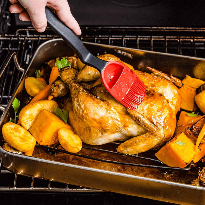 Red silicone basting brush with ergonomic black handle applying glaze to roasted chicken and vegetables in oven-safe pan, featured in Kitchen & Food, Cooking and Baking Helpers, Grilling, and Kitchen Gadgets collections.