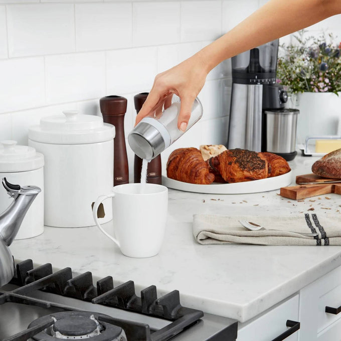 Hand pouring sugar from a clear glass dispenser with stainless steel lid into a white mug on a kitchen counter, surrounded by baked croissants, salt and pepper mills, and white canisters, highlighted in Kitchen & Food, Kitchen Gadgets, Kitchen Accessories, Storage and Serving, Best Sellers, and Promo Eligible collections.