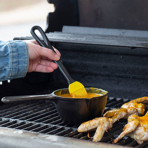 Black cast iron melting pot with long handle heating on grill, accompanied by a yellow silicone basting brush, ideal for Kitchen & Food, Cookware, Cast Iron, and Best Sellers collections.