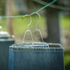 Two pairs of rust-resistant steel pants stretchers with adjustable clamps holding jeans on hangers, drying outdoors on a clothesline in natural light, ideal for laundry supplies.