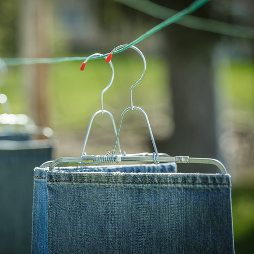 Two pairs of rust-resistant steel pants stretchers with adjustable clamps holding jeans on hangers, drying outdoors on a clothesline in natural light, ideal for laundry supplies.
