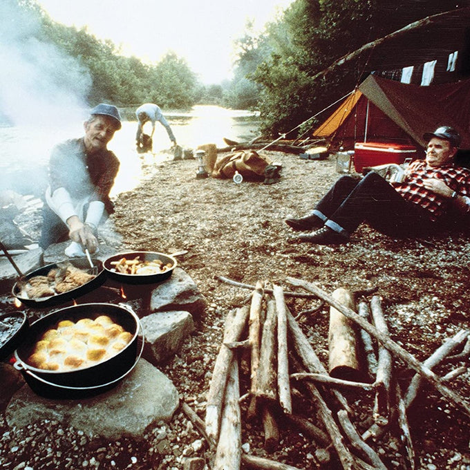 Outdoor camping scene by a riverside with a campfire cooking multiple cast iron pots including a dutch oven, surrounded by firewood and a pitched tent, evoking Farm, Garden & Tool and Camping collections.