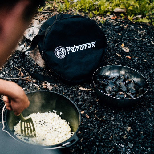 Black round carrying bag with Petromax logo placed outdoors on gravel near a cast iron pot cooking diced onions and a metal bowl filled with mussels, suited for Farm, Garden & Tool, Camping, Best Sellers, and Promo Eligible collections.