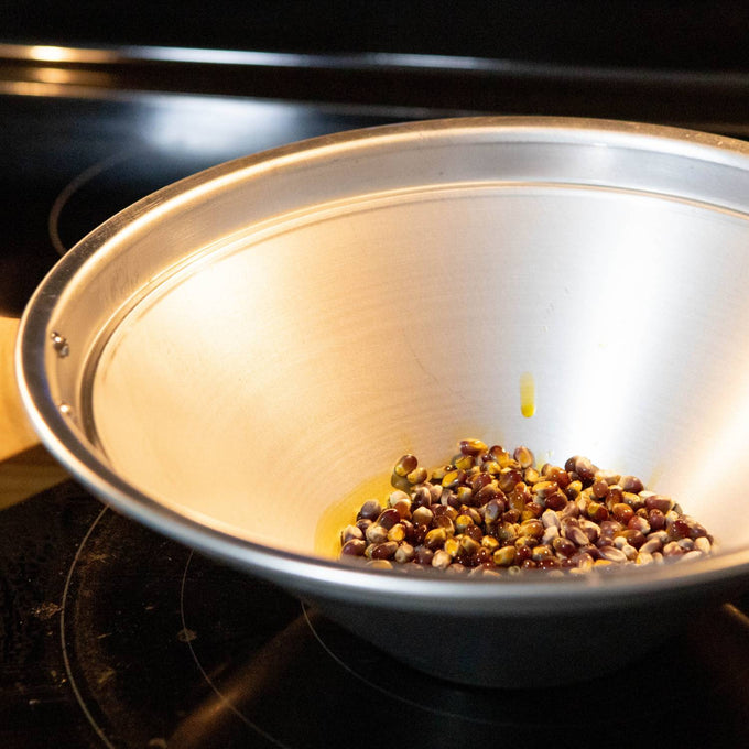 Conical aluminum stovetop popcorn popper bowl with a small amount of unpopped kernels, displayed on a black electric stove, featured in Kitchen & Food, Cookware, and Best Sellers collections.
