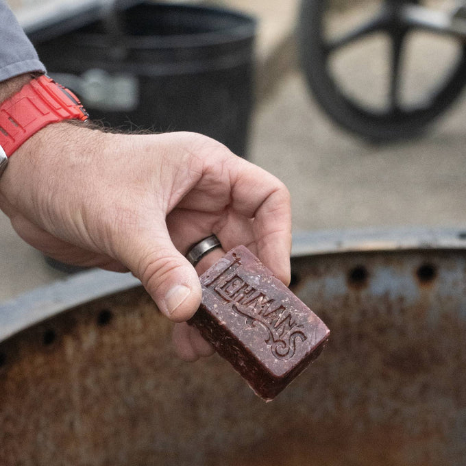Handcrafted reddish-brown fire starter with Lehman’s logo, held above a metal fire ring, designed for easy, non-toxic fire building in wood stoves and campfires, ideal for Farm, Garden & Tool and Camping collections.