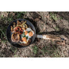 Wrought iron pan with cooked eggs, bell pepper slices, potatoes, and meat, featuring a wooden handle with engraved logo, shown outdoors on dry grass, suitable for camping and cookware collections.