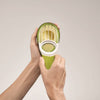 Hands holding a green 3-in-1 avocado tool with plastic slicing blades and a pitting section, shown cutting and preparing an avocado, suitable for kitchen and food prep use.