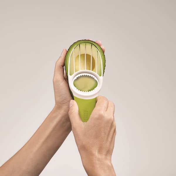 Hands holding a green 3-in-1 avocado tool with plastic slicing blades and a pitting section, shown cutting and preparing an avocado, suitable for kitchen and food prep use.