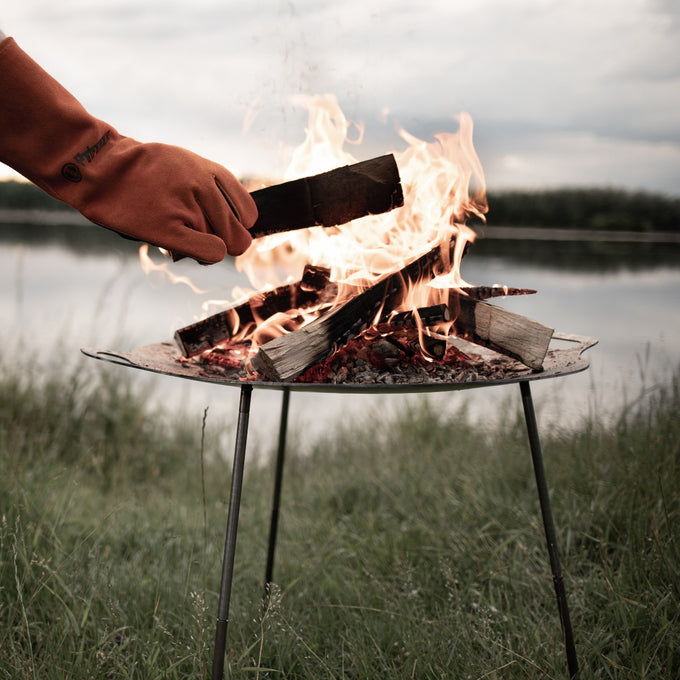 Gloved hand placing firewood onto a steel fire bowl with three legs, flames rising against a lake backdrop, perfect for Farm, Garden & Tool, Camping, Best Sellers, and Promo Eligible collections.