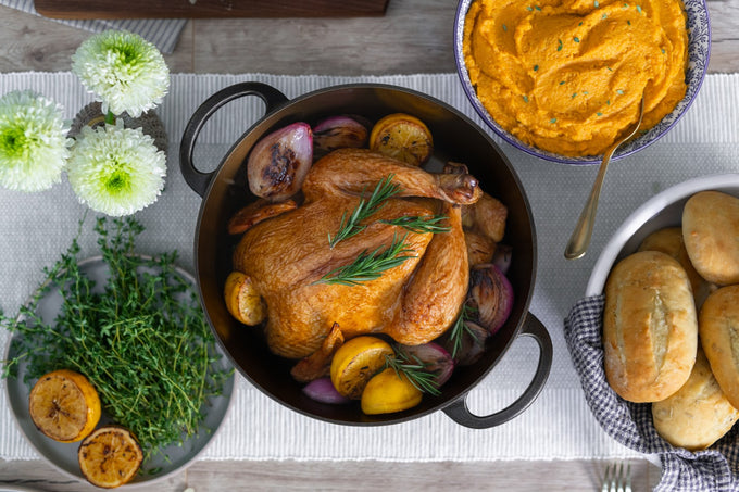 Handcrafted cast iron Dutch oven with roasted whole chicken, lemon halves, and herbs, surrounded by fresh thyme, green flowers, mashed sweet potatoes, and freshly baked rolls for Kitchen & Food, Camping, and USA Made collections.