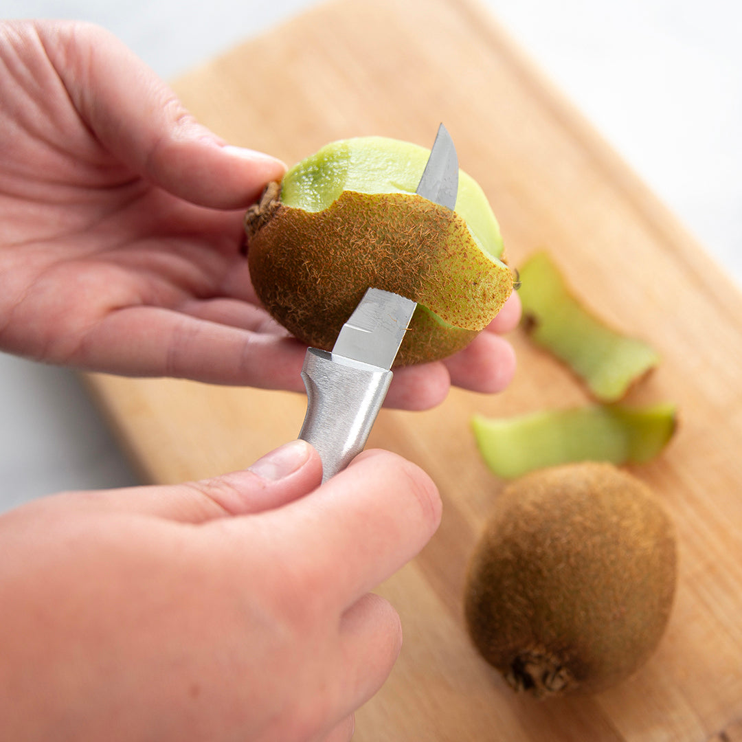 Handcrafted Rada Granny Parer with a high carbon surgical stainless steel blade and solid aluminum handle, expertly peeling a kiwi on a wooden cutting board, made in USA, featured in Farm, Garden & Tool, Kitchen & Food, and Knives & Axes collections.