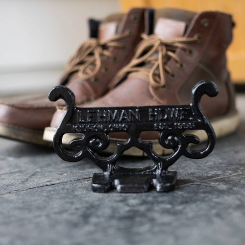 Close-up of a handcrafted black cast aluminum boot scraper with vintage scroll design, made in USA and Amish crafted, placed on a stone floor with brown leather boots in the background.