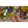 Hands arranging fresh yellow herbs on rustic wooden table surrounded by glass bottles, mortar and pestle, and colorful wildflowers in natural wellness and health book setting