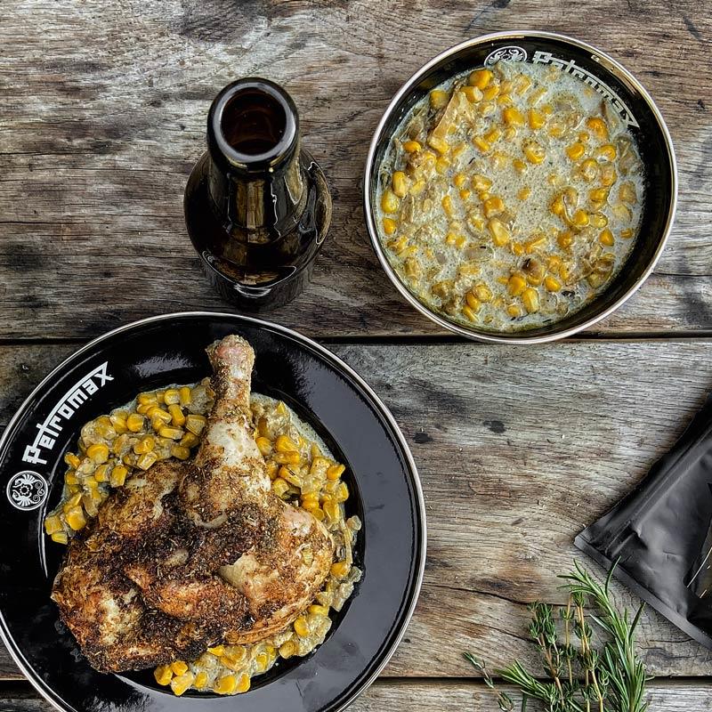 Black enamel bowl with Petromax logo holding cooked chicken leg and creamed corn, paired with smaller matching bowl of creamed corn and a dark glass bottle on rustic wooden surface, suitable for Farm, Garden & Tool, Kitchen & Food, Camping, Dinnerware, Best Sellers, Promo Eligible collections.