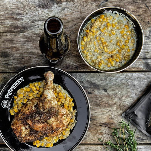 Black enamel bowl with Petromax logo holding cooked chicken leg and creamed corn, paired with smaller matching bowl of creamed corn and a dark glass bottle on rustic wooden surface, suitable for Farm, Garden & Tool, Kitchen & Food, Camping, Dinnerware, Best Sellers, Promo Eligible collections.