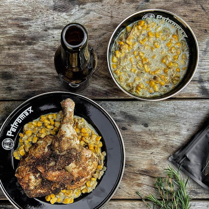 Black enamel bowl with Petromax logo holding cooked chicken leg and creamed corn, paired with smaller matching bowl of creamed corn and a dark glass bottle on rustic wooden surface, suitable for Farm, Garden & Tool, Kitchen & Food, Camping, Dinnerware, Best Sellers, Promo Eligible collections.