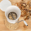 Traditional nut chopper with a white plastic hopper and glass jar, stainless steel blades, and a hand crank, shown alongside shelled walnuts on a wooden surface, ideal for Kitchen & Food and Food Prep collections.