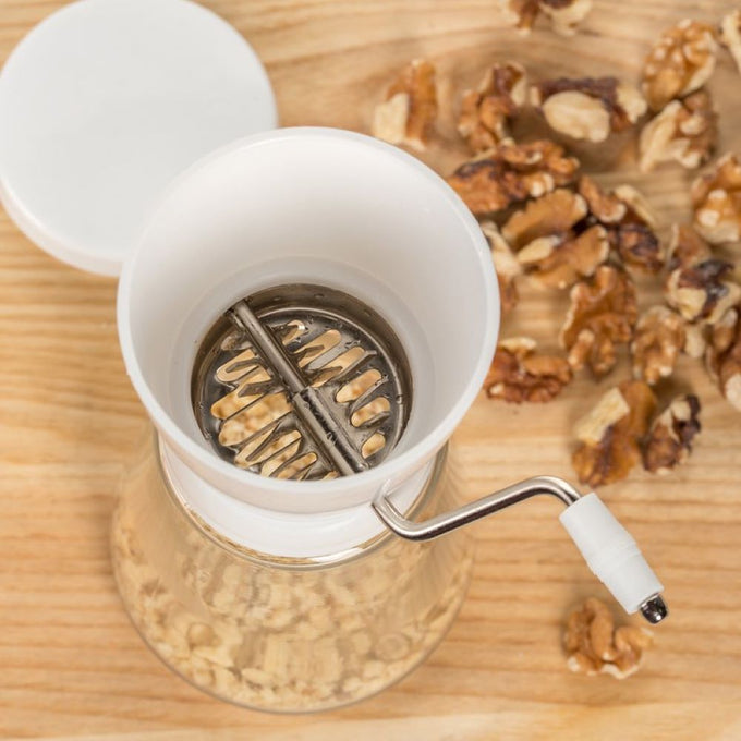 Traditional nut chopper with a white plastic hopper and glass jar, stainless steel blades, and a hand crank, shown alongside shelled walnuts on a wooden surface, ideal for Kitchen & Food and Food Prep collections.