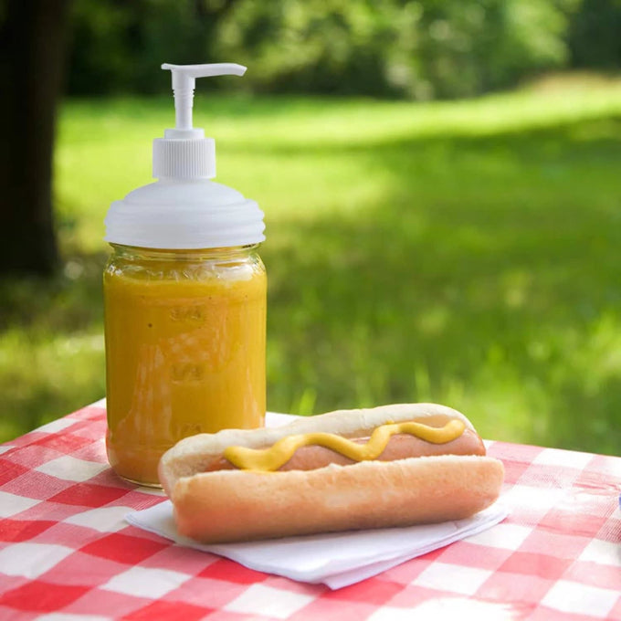 White pump lid fitted on a glass jar filled with yellow condiment, next to a mustard-topped hot dog on a checkered picnic tablecloth, showcasing an eco-friendly kitchen accessory ideal for Health & Wellness and Home & Decor collections.
