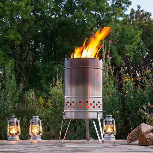 Stainless steel outdoor cooking stove with vibrant flames burning inside, surrounded by four vintage-style oil lanterns on a stone patio, ideal for camping and garden use.