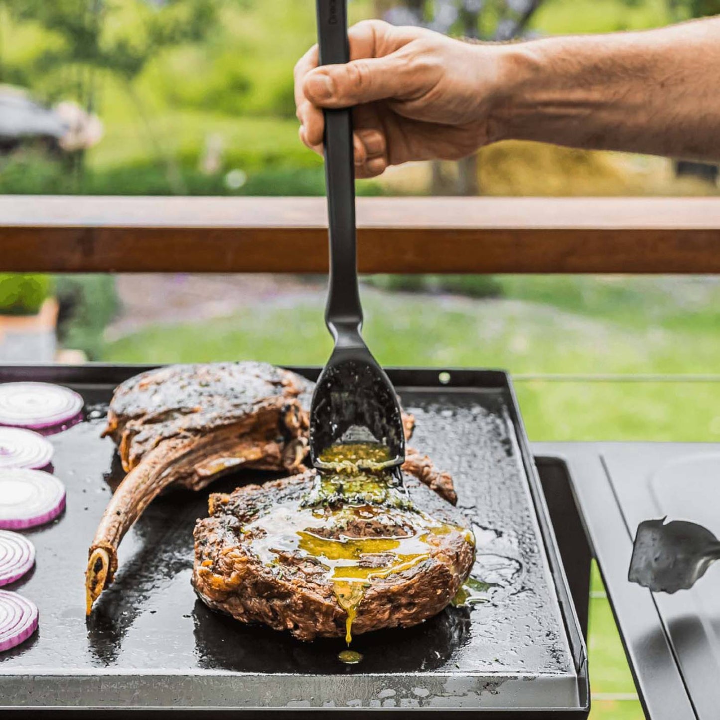 Hand holding a black BBQ Brizzle basting brush with a deep scooping head, applying sauce over grilled steak on a flat-top grill, ideal for Kitchen Gadgets and Grilling collections.
