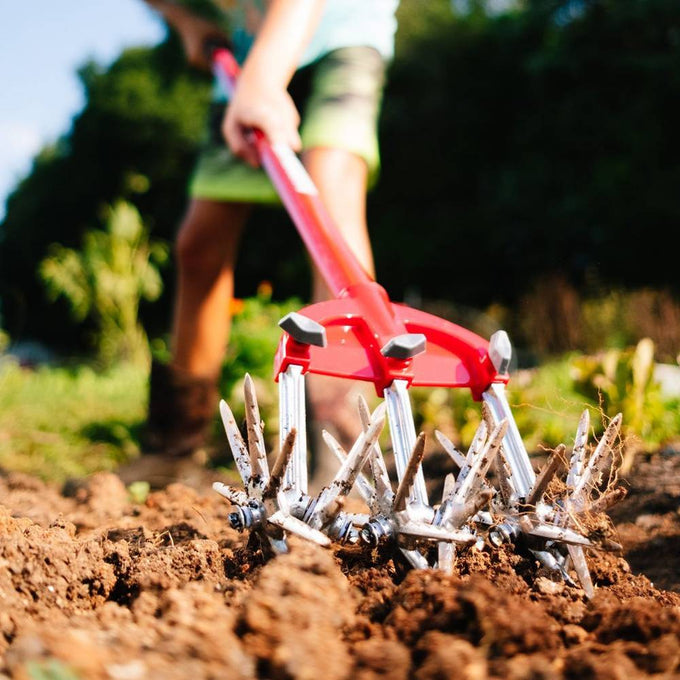 Close-up of a person using the Original Garden Weasel with rust-proof serrated steel tines and a red comfort-grip handle to loosen soil and remove weeds, featured in Farm, Garden & Tool, Digging and Cultivating, Garden Accessories, Best Sellers, and Promo Eligible collections.