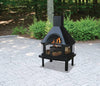Black wood burning outdoor firehouse with chimney and mesh protective sides, set on sturdy legs, shown burning firewood on a textured stone patio surrounded by greenery, featured in Stoves and Grilling collections.