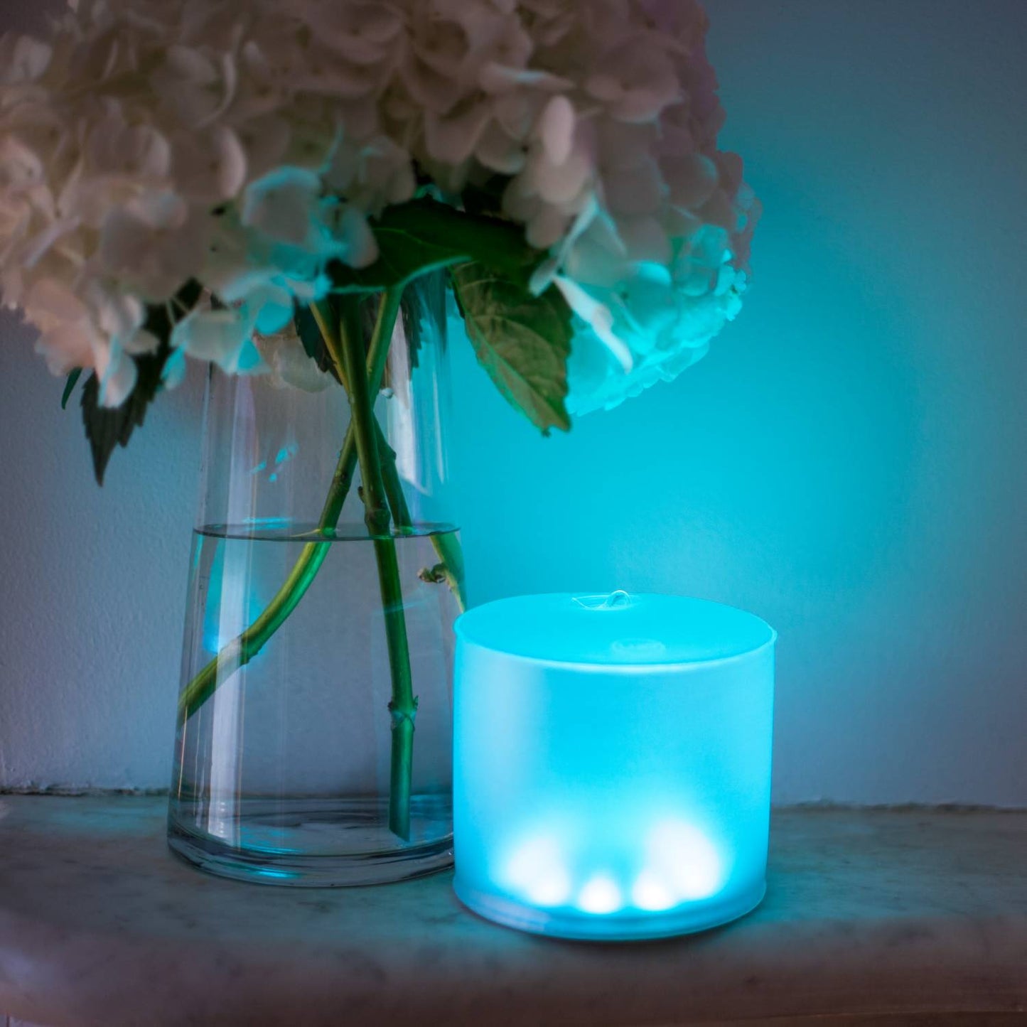 Soft blue glowing inflatable Luci Color Essence solar light casting a calming ambiance beside a glass vase with white hydrangea blooms, perfect for cozy indoor lighting.