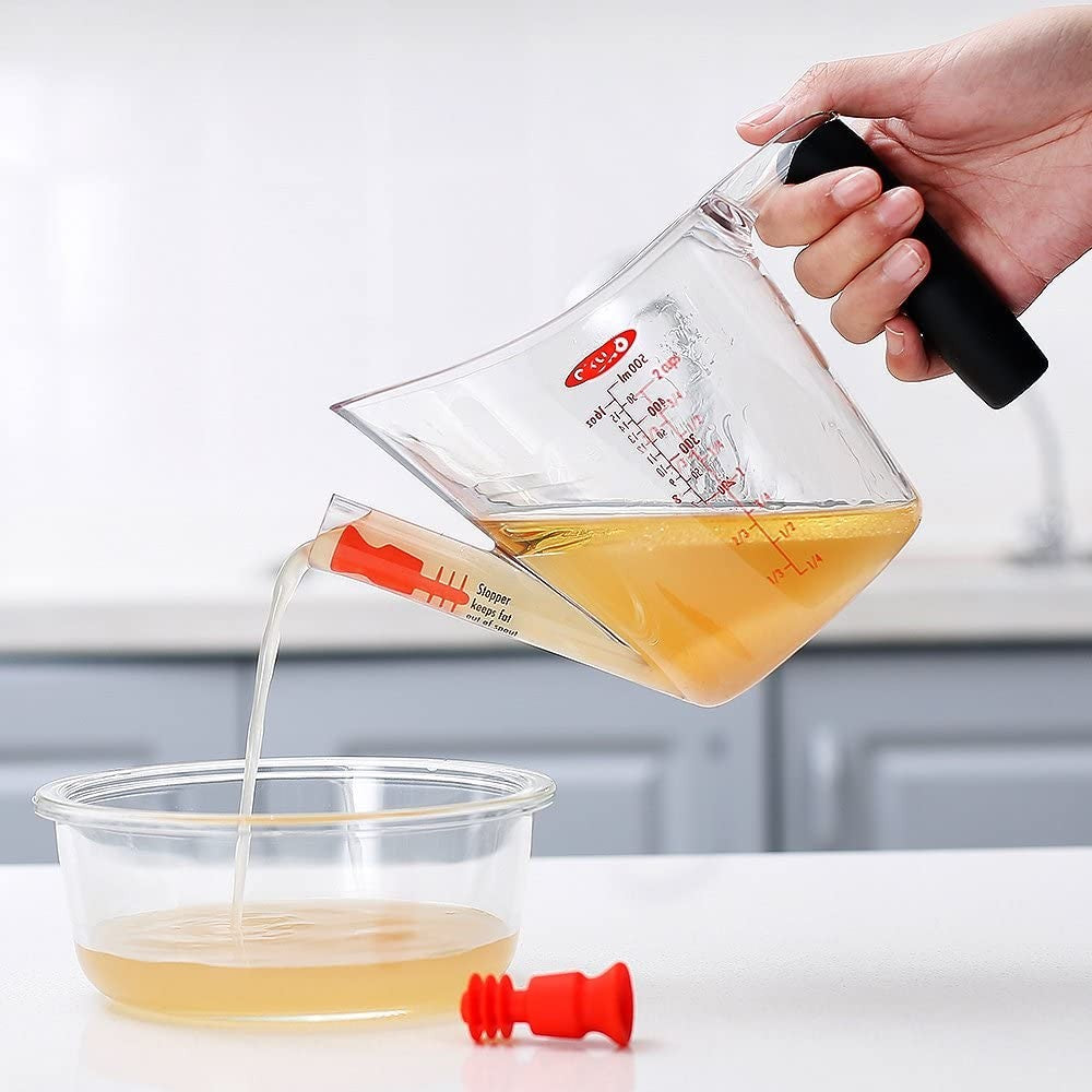 Clear fat separator with soft black grip handle, red stopper beside it, and angled spout pouring clear liquid into a bowl, ideal for Kitchen & Food, Kitchen Gadgets, and Cooking Helpers collections.