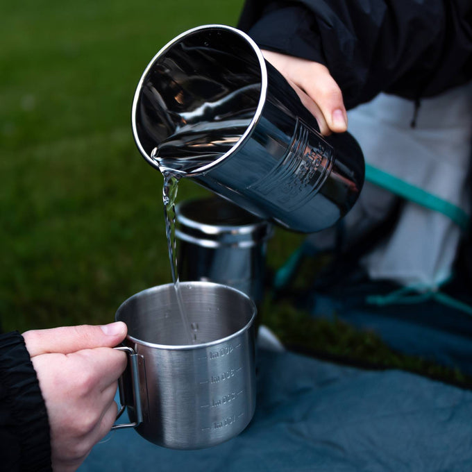 Close-up of two hands pouring water from one polished stainless steel camping cup into another on a blue outdoor blanket, highlighting durable and portable drinkware perfect for camping and emergency supplies.