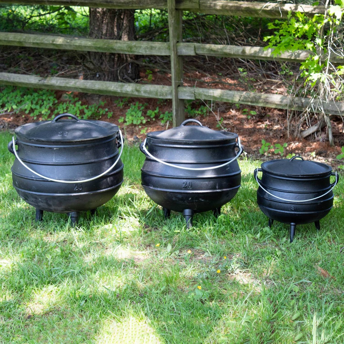 Three handcrafted cast iron campfire kettles in small, medium, and large sizes with round bodies, fitted lids, wire handles, and three legs, set outdoors on grass near a wooden fence, ideal for Farm, Garden & Tool, Camping, Best Sellers, and Promo Eligible collections.