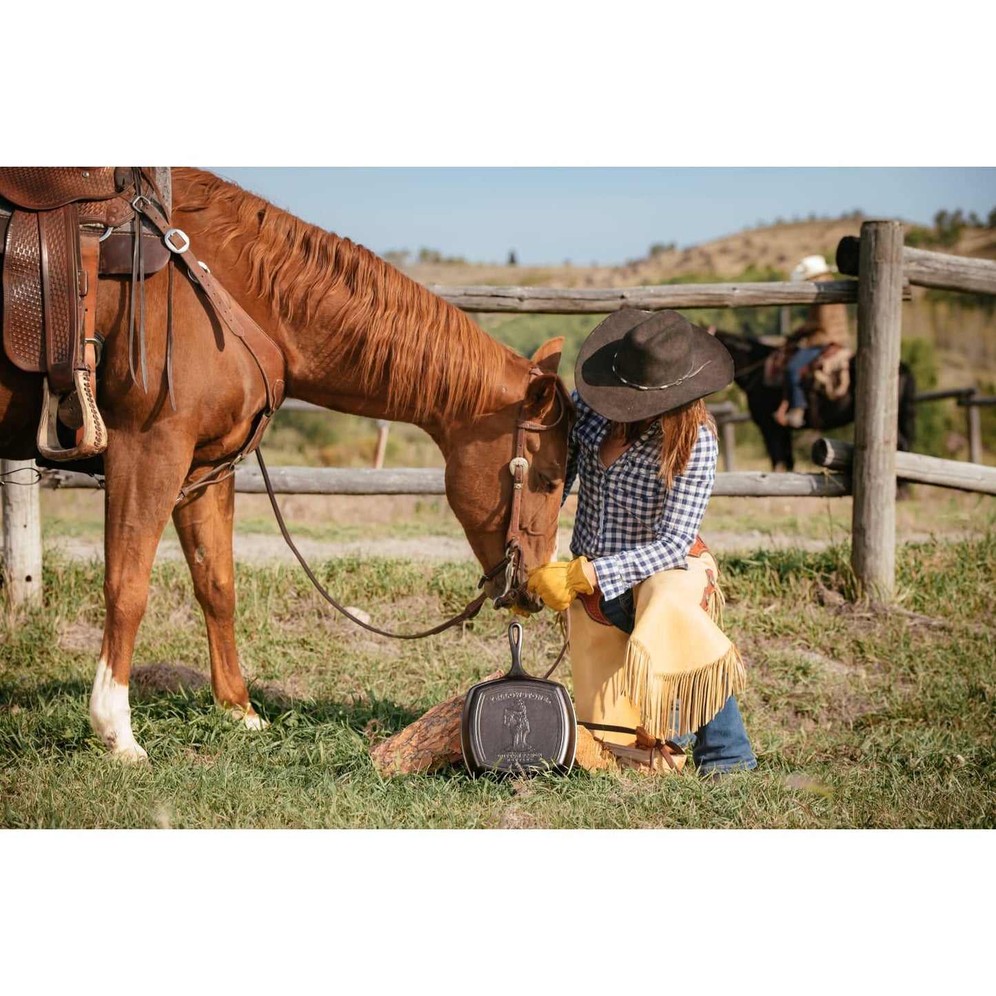 Woman wearing a wide-brimmed hat, plaid shirt, and leather chaps kneeling next to a brown horse near a wooden fence, holding a handcrafted cast iron square grill pan made in USA, suited for Kitchen & Food, Cookware, Cast Iron, and USA Made collections.