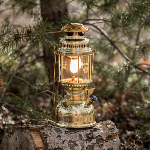 Polished brass kerosene pressure lamp glowing warmly on a log in a forest setting, designed for reliable lighting in camping, farm, garden, and emergency situations.
