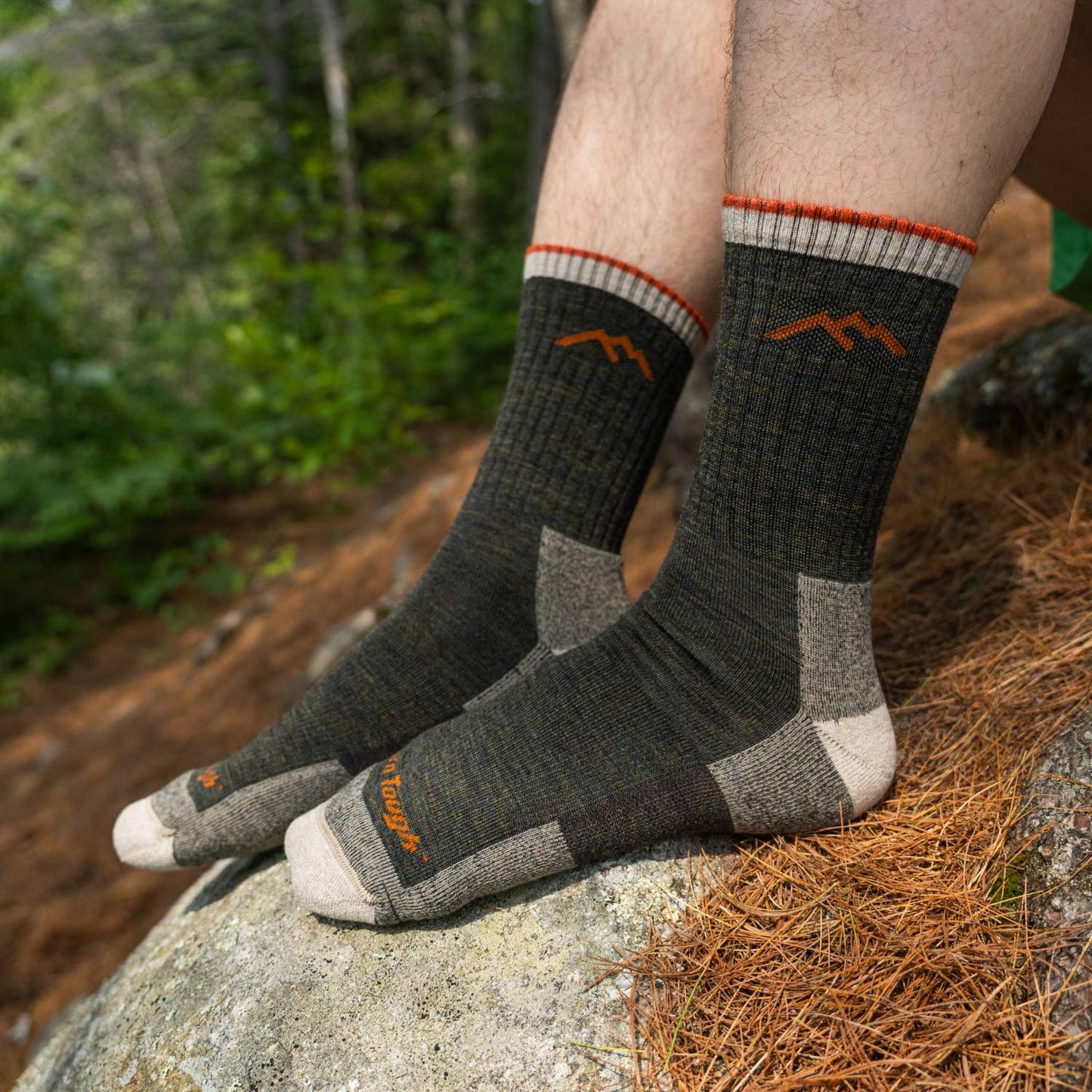 Pair of midweight men's hiking socks in dark green and gray with orange accents, worn outdoors on a rock in a forest setting, perfect for camping and wellness collections.