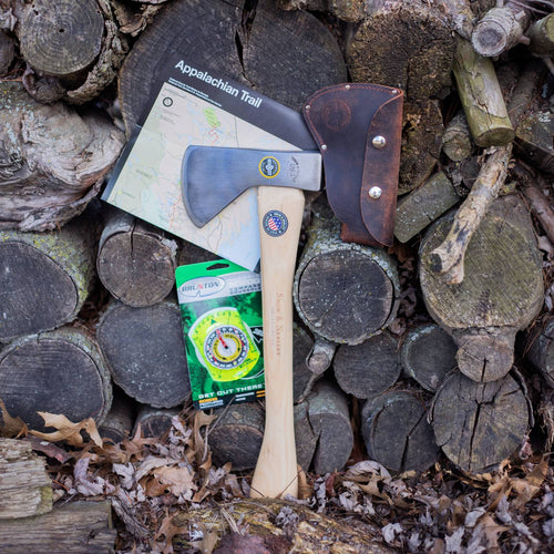 Lightweight handcrafted kindling axe with a wooden handle and leather sheath resting against a stack of firewood, alongside an Appalachian Trail map and survival compass, perfect for camping and outdoor use.