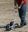 Person using a handcrafted, made in USA garden seeder with metal frame and large black wheels to plant evenly spaced rows, featured in Farm, Garden & Tool and Seeds and Planting collections.