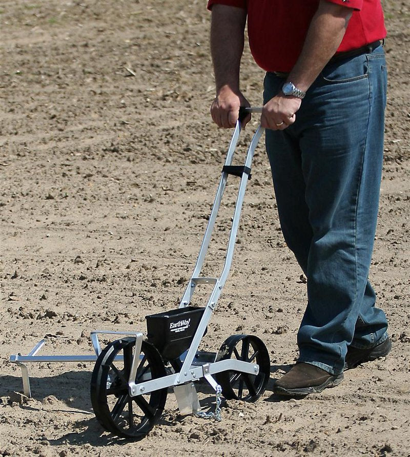 Person using a handcrafted, made in USA garden seeder with metal frame and large black wheels to plant evenly spaced rows, featured in Farm, Garden & Tool and Seeds and Planting collections.