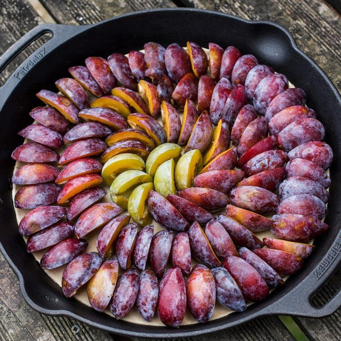 Handcrafted cast iron fire skillet with two sturdy side handles, filled with neatly arranged sliced plums in vibrant red and yellow hues, perfect for camping and kitchen use.
