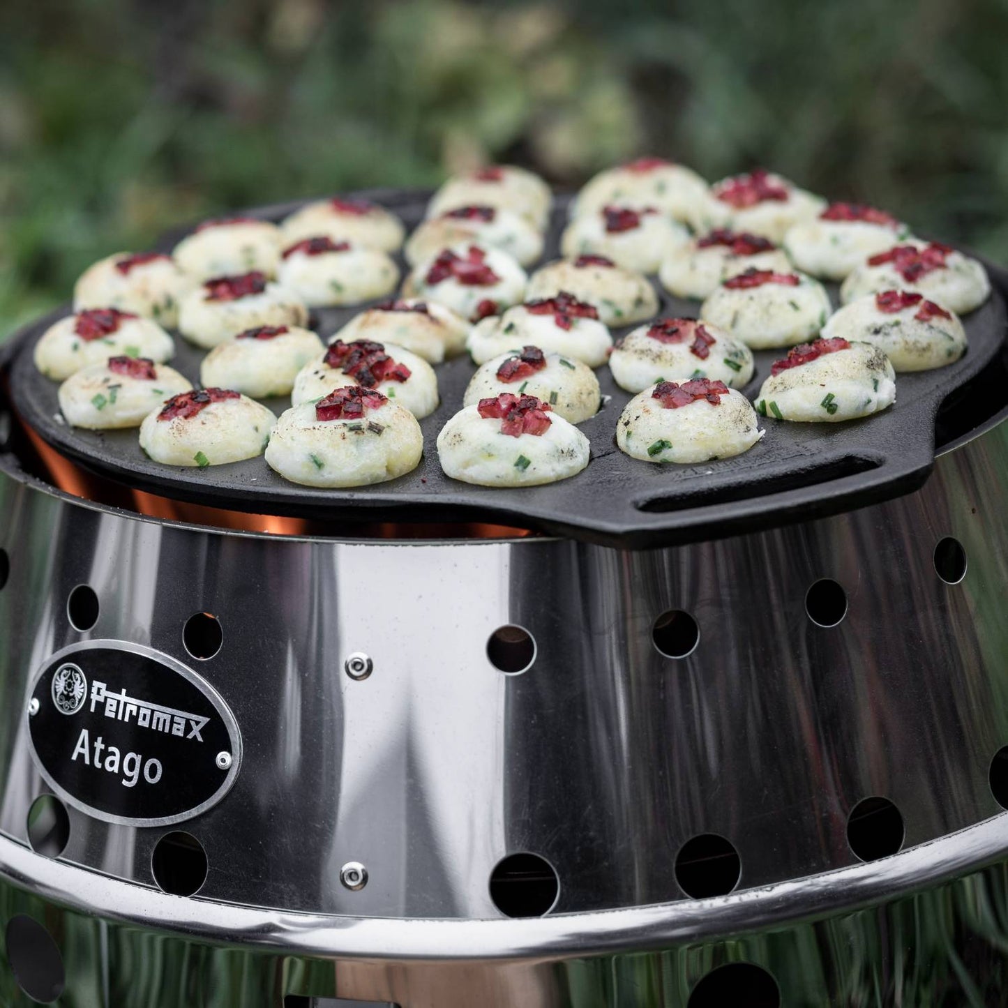 Round cast iron pan with 30 mini poffertjes cooking on a stainless steel outdoor stove, handcrafted for use over open fires in camping and kitchen cookware collections.