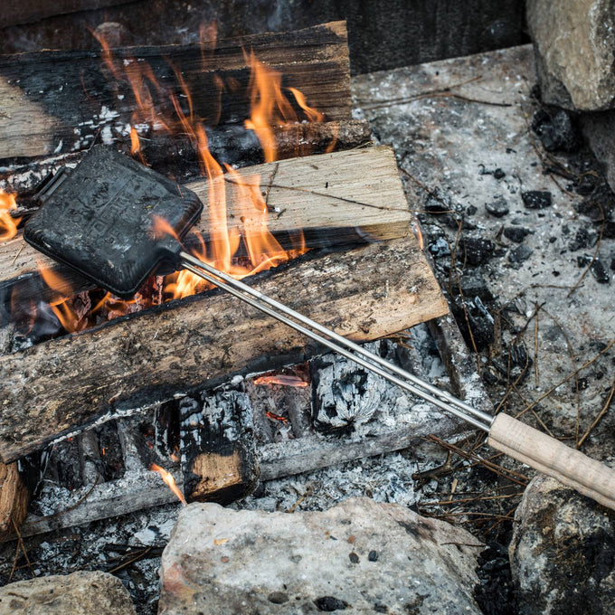 Cast iron sandwich iron with wooden handle held over an open campfire, perfect for toasting sandwiches while camping, farmhouse, or outdoor cooking in Farm, Garden & Tool and Camping collections.