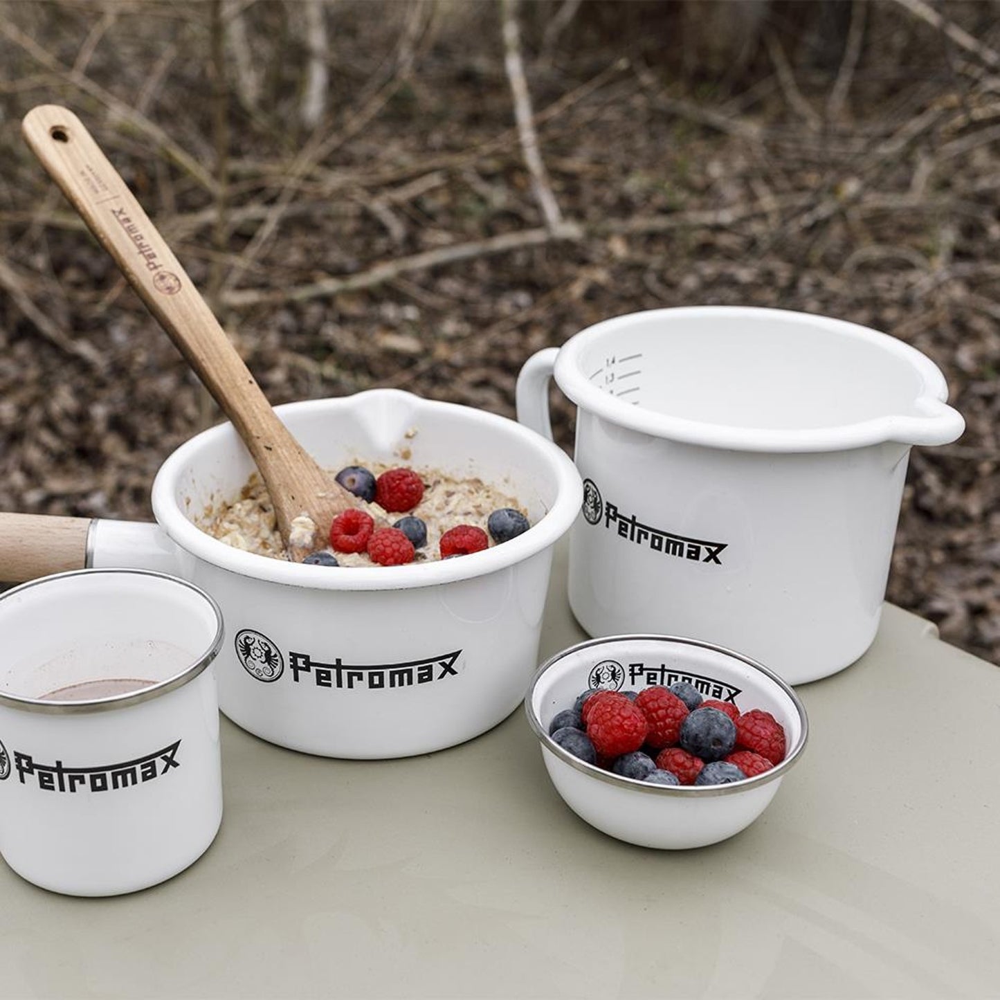 Set of white enameled steel cookware including a milk pot with spout, a bowl of oatmeal with berries and wooden spoon, a small bowl with berries, and a cup, handcrafted for Farm, Garden & Tool, Kitchen & Food, Camping, Cookware, Pots and Pans, Best Sellers, and Promo Eligible collections.